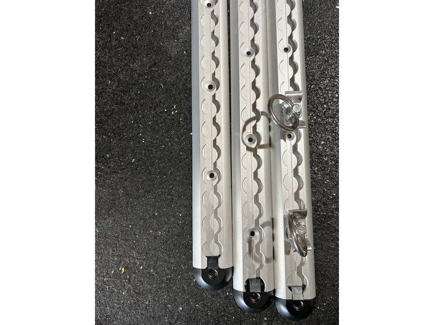 Drei nebeneinanderliegende eloxierte Aluminiumprofile mit gewellter Nut, Befestigungsbohrungen und zwei eingehängten Ösen, auf dunklem Untergrund.